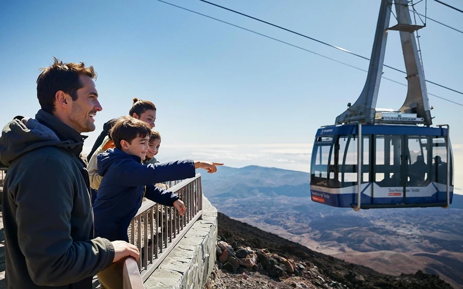 A family at a viewpoint railing watching a cable car cabin climb the mountain